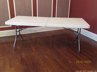 White folding table shown from a front angle with legs visible, placed on wood floor near wall.