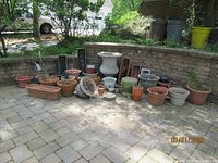 Wide view of a large assortment of garden pots in various materials and sizes arranged outdoors on a stone patio area with greenery in the background