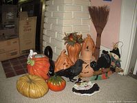 Photo showing multiple Halloween decorations including ceramic pumpkins with faces, black bird figurine, scarecrow doll, black cat trick or treat sign, orange pumpkin-shaped pitcher with black cat handle, and a broom leaning against a white brick wall.