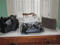 Four handbags on marble surface: black double tassel shoulder bag, white and tan structured handbag with tags, black duffle style leather shoulder bag, taupe leather classic shoulder bag.