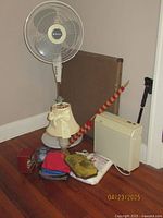 Photo of vintage Holmes Stand Fan, Hunter desk fan, electric lamp, walking stick, card table and various household items arranged on floor.