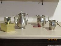 Group of four pewter items including 3 pitchers and 1 cup, arranged on flat surface with neutral background