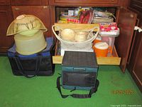Photo showing various picnic items including two soft-sided insulated cooler bags, vintage cake carrier, woven basket, paper products and plastic containers in a cabinet.