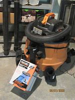 Orange and black Ridgid shop vacuum with coiled flexible hose, box of replacement filters, and several vacuum attachments around it.
