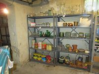 Two gray metal shelving units in basement setting. Shelves contain assorted household items such as glassware, jars, pottery, and canned goods. Photo taken to show shelving units, contents not part of sale.