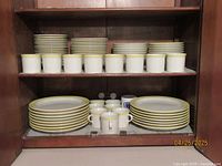 Stone Crest dinnerware set on shelves showing stacks of dinner plates, mugs, bowls, small plates, and saucers, all white with yellow and gold rims.