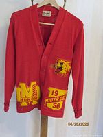 Front view of red knit school sweater with three button closure and chenille patches 'M Honor' and '19 Mater Dei 56' on lower front.