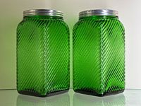 Front and angled view of the two green swirl glass jars with metal lids together, showing the diagonal swirl texture and shiny bright green color of the glass.