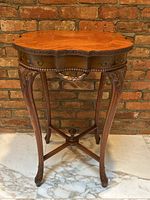 Front three-quarter view of wooden side table against brick wall showing octagonal top, carved apron, and legs joined by X-stretcher