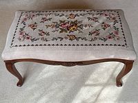 Top view of the footstool showing floral needlepoint upholstery and carved wood frame with cabriole legs