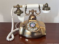 Front angled view of antique style rotary dial telephone showing gold-tone body, white handset, coiled cord, and rotary dial with image and alphanumeric numbers.