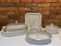 Five white ceramic CorningWare baking and casserole dishes with clear glass lids, displayed on a white table against a brick wall, showing different shapes and sizes including an oval dish and a lasagna pan.