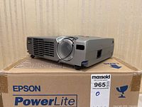 Angled front view of Epson PowerLite projector seated on its original box, lens cap attached