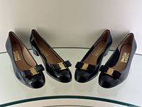 Two pairs of black Ferragamo pumps side by side on glass shelf