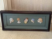 Straight-on shot of the framed set showing six porcelain doll head silhouettes on green linen.