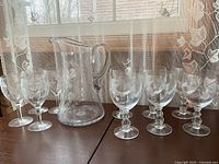 Full set showing one glass pitcher and ten stemmed glasses arranged in two groups with etched butterfly and embossed ladybug designs, on a brown surface with lace curtain background.