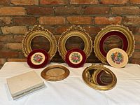 Full display of the lot showing three ornate round gold frames with needlepoint inserts, one oval gold frame containing a mirror, one extra oval frame, and two needlepoint artworks.