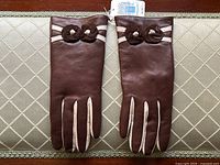 Top side view of the brown leather gloves with cream detailing and decorative rosettes, laid flat.