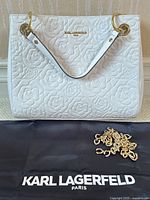 Front view of white leather handbag with embossed rose pattern, dust bag beneath and gold chain strap laid in front