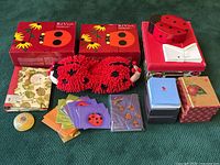 Overview of the ladybug-themed lot showing slippers, notecard sets, stacked boxes, and assorted stationery items arranged on a green carpet, highlighting the red and black ladybug motif.