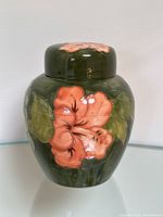 Front and side view of a glazed ceramic ginger jar with pink hibiscus flowers and green leaves on dark green background.