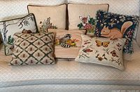 Six needlepoint and tapestry pillows on a white couch showing a woodland animal and nature theme.