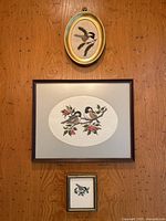 Photo showing all three framed needlepoint artworks hanging on wood wall: one large rectangular frame with two chickadees and red berries, one small oval frame with single chickadee, and one small rectangular frame with single chickadee.
