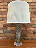 Front view of tall lamp with white fabric shade and reverse painted art glass base against brick wall.