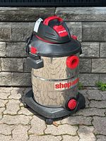Shop-Vac 12 gallon wet/dry vacuum with stainless steel body, black and red top and base, placed outdoors on a stone tile surface in front of a stone wall.