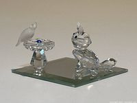 Swarovski crystal figurines of a cat and bird on a bird bath on a mirrored base, showing clear faceted details and black eye accents on cat, blue and frosted crystal on bird bath and bird.