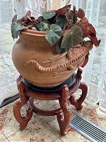 Front angle of the terracotta planter with lizard relief on the side, showing plant leaves and wooden stand below.