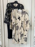 Two vintage 80s Art Deco style sheath dresses hanging on hangers, one cream-colored and one black, detailed print and necklines visible