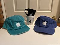 Pair of green and blue Apple logo cotton ball caps with stacked black and white Apple logo mugs behind them