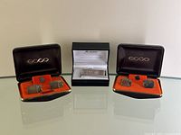 Full view of two sets of Ecco cufflinks and lapel pins in black cases with orange interiors, flanking a black box containing the gold plated money clip.
