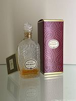 Clear glass bottle with intricate embossed designs and amber liquid, placed beside original purple box with embossed pattern.
