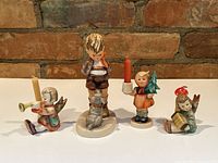 Front view of all four vintage Hummel figurines including one standing boy and three smaller seated children candle holders arranged on a white surface against a brick background.