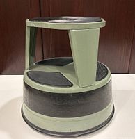 Side angle showing three-step stool with avocado green frame and black rubber tread pads, placed on a white surface against a wooden background.