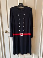 Full front view of the navy blue knit dress with red belt and white buttons design, hanging on a hanger in front of a door.