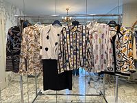 Wide view of six clothing pieces hung on a rack showing various prints including perfume bottles, dress forms, stripes, and chains.