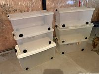 Two stacks of three clear plastic storage bins each with white lids and black wheels, placed against a wall on a concrete floor.