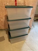 Stack of four clear plastic storage bins with green lids shown in unfinished basement area.