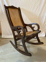 Side view of antique dark stained wood rocking chair showing curved armrest and woven wicker seat and back.