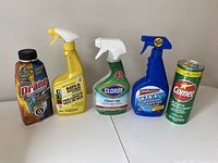 Five unopened household cleaning products displayed on white surface: Drano, CLR, Clorox, Scotchgard, Comet.