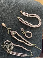 Four pink pearl-like jewelry pieces laid out on black fabric, including 3 necklaces with different pendants and 1 matching bracelet.