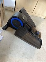 Black and blue Cubic under desk elliptical exerciser resting on floor tiles next to cardboard box and paperwork.
