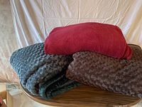 Photo showing three folded blankets in red, brown, and green colors stacked on a round wooden table.