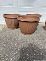 Three large brown plastic flower pots with ridged design, placed outside on concrete, in front of a white garage door.