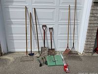Full view of all garden tools including rakes, shovel, spade, hand tools, kneeling pads and bamboo stakes arranged outdoors in front of white garage door.