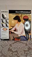 Front view of Homedics Back Masseur original packaging with female model using the device, displaying core features like 10 programmable massage motors, heat function, and remote control.