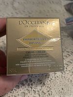 Four images showing the front and back of the gold-colored boxes of L'Occitane Immortelle Divine Advanced Youth Face Care Cream 50ml boxed products.